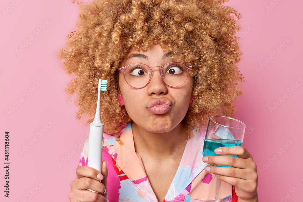 Photo of curly haired young woman makes funny grimace pouts lips holds electric brush and glass of mouthwash takes care of dental hygiene isolated over pink background. Morning routine concept - obrazy, fototapety, plakaty 