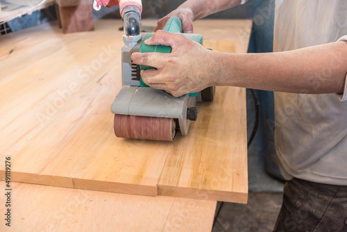 Using a hand-held sanding machine or belt sander to level the surface of a sheet of plywood for a cabinet, table or other furniture at a workshop.