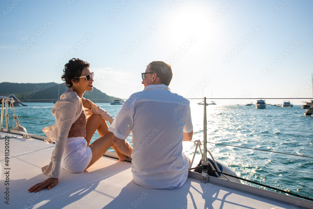 Caucasian couple enjoy outdoor luxury party drinking champagne with talking together while catamaran boat sailing at sunset. Man and woman relax with outdoor lifestyle on summer travel vacation