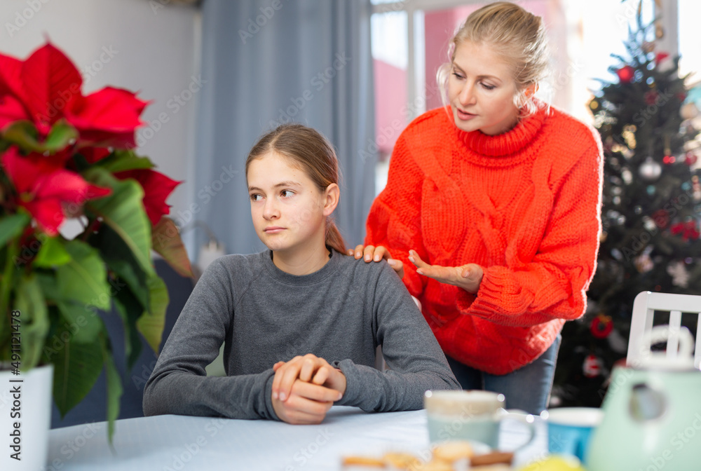 Upset teenager girl after quarrel with her mother in domestic interior before xmas, mother consoles daughter