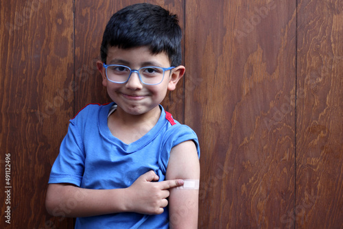 Little 6-year-old Latino boy with glasses and a blue shirt shows his arm with a bandage because he has just been vaccinated against Covid-19 in the new normality due to the Coronavirus pandemic
 photo