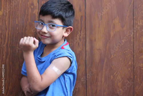 Little 6-year-old Latino boy with glasses and a blue shirt shows his arm with a bandage because he has just been vaccinated against Covid-19 in the new normality due to the Coronavirus pandemic 