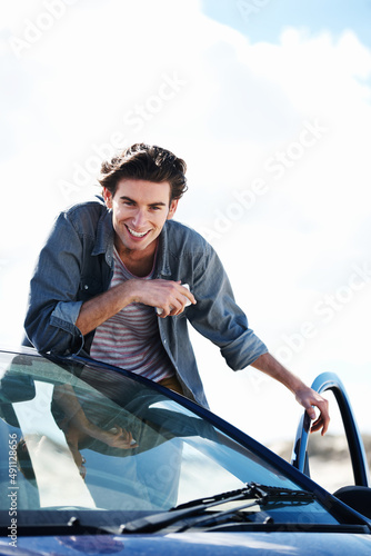 Ready to hit the road. Happy young man holding a cellphone and leaning on the roof of his car while smiling at the camera.
