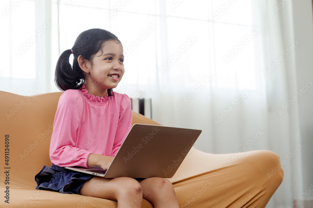 An adorable Asian girl wearing a pink shirt is happily sitting at her laptop notebook computer on the sofa in the living room of her home.