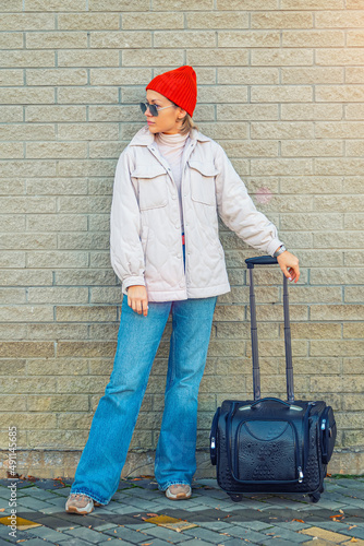 A young woman in casual clothes and with a suitcase poses at the airport at the train station against the background of a brick wall in sunny weather