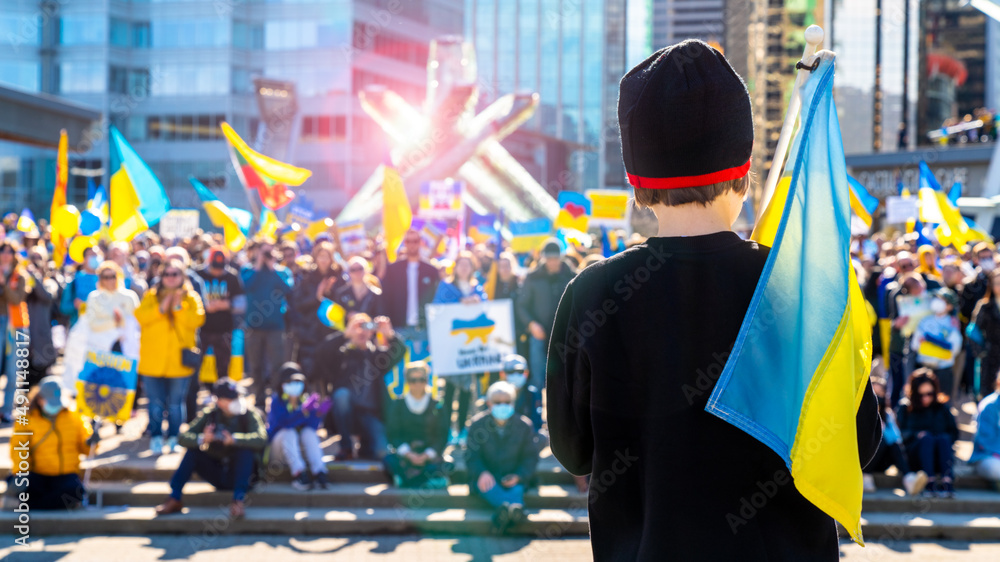 Young boy standing in front of a crowd at a protest against the Russian invasion in Ukraine. Child holding on his shoulder a Ukrainian flag while facing the gathered people for a peaceful protest. - obrazy, fototapety, plakaty 