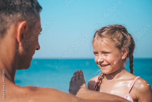 Father applying protective cream to her daughter's face at the beach. Man hand holding sunscreen lotion on baby face. Cute little girl with a sun block by the sea . Copy space. © smile35