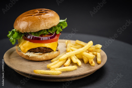 Tasty homemade beef burger with fresh ingredients served on little wooden cutting board with french fries on dark background, close-up view..