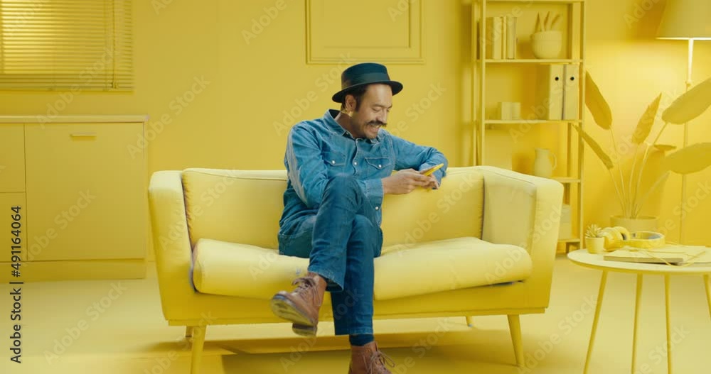 Happy Hispanic man typing a message on the mobile phone while smiling and sitting on a sofa in the studio with monochrome yellow interior background. Man sitting on couch while using cellphne Happy Hispanic man typing a message on the mobile phone while smiling and sitting on a sofa in the studio with monochrome yellow interior background. Man sitting on couch while using cellphne