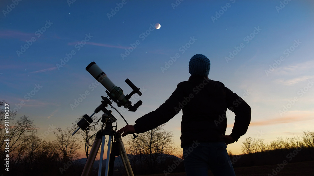 Silhouette of a man, telescope and countryside under evening skies.