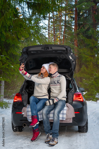 A young couple taking selfies with a phone and enjoying beautiful winter weather in the open trunk of a car in the winter snowy forest. Happy caucasian family enjoying the romantic outdoor date. © Aimur