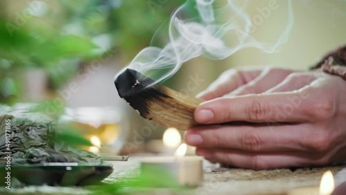 Woman hands burning Palo Santo, before ritual on the table with candles and green plants. Smoke of smudging treats pain and stress, clears negative energy and meditation wooden stick