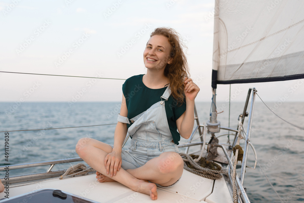 An attractive smiling woman is looking at the camera and touching her red hair while sitting on a yacht