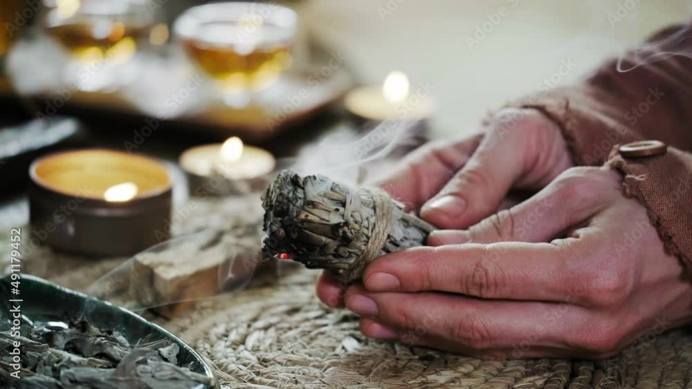 Woman hands burning white sage, before ritual on the table with candles and green plants. Smoke of smudging treats pain and stress, clear negative energy and meditation Woman hands burning white sage, before ritual on the table with candles and green plants. Smoke of smudging treats pain and stress, clear negative energy and meditation