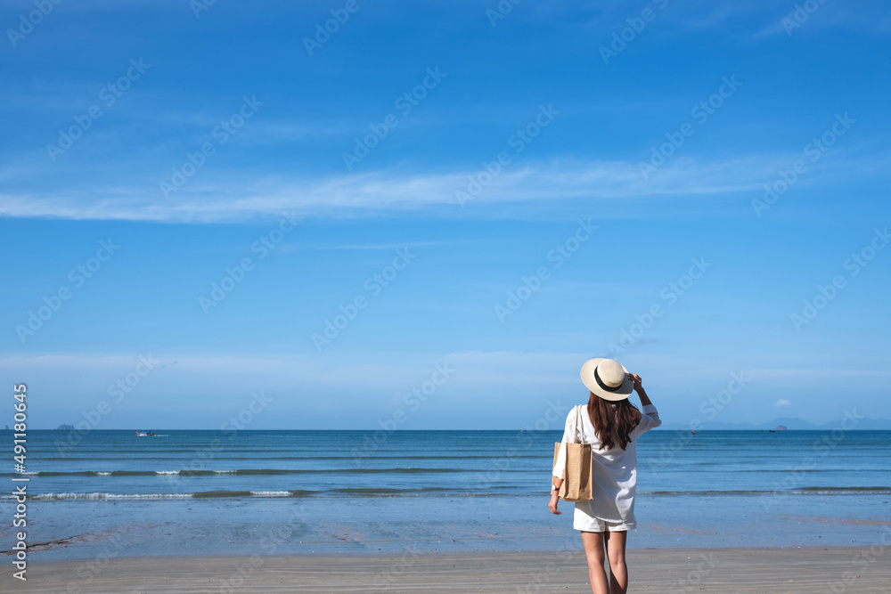 Rear view image of a young woman with hat and bag walking on the beach with blue sky background