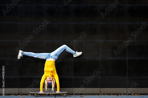 Athetic young woman upside down on a skateboard, black wall background, yellow shirt and blue jeans, generation z female crazy lifestyle photo