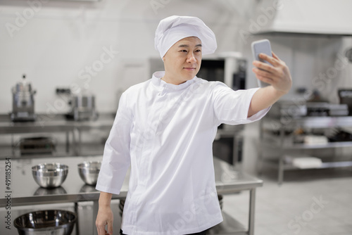 Handsome asian chef cook in white uniform takes selfie photo on phone or makes video blog for social networks while working in the restaurant kitchen. Concept of social activity at job