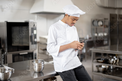 Handsome chef in uniform standing with phone in professional restaurant kitchen. Latin guy using phone on work at restaurant