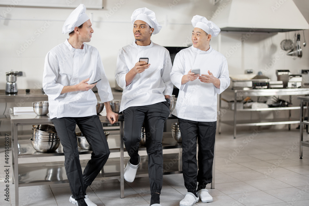 Three multiracial chef cooks talk while standing with smart phones during a break at the restaurant kitchen. Asian, Latin and European men in white uniform during a break at work
