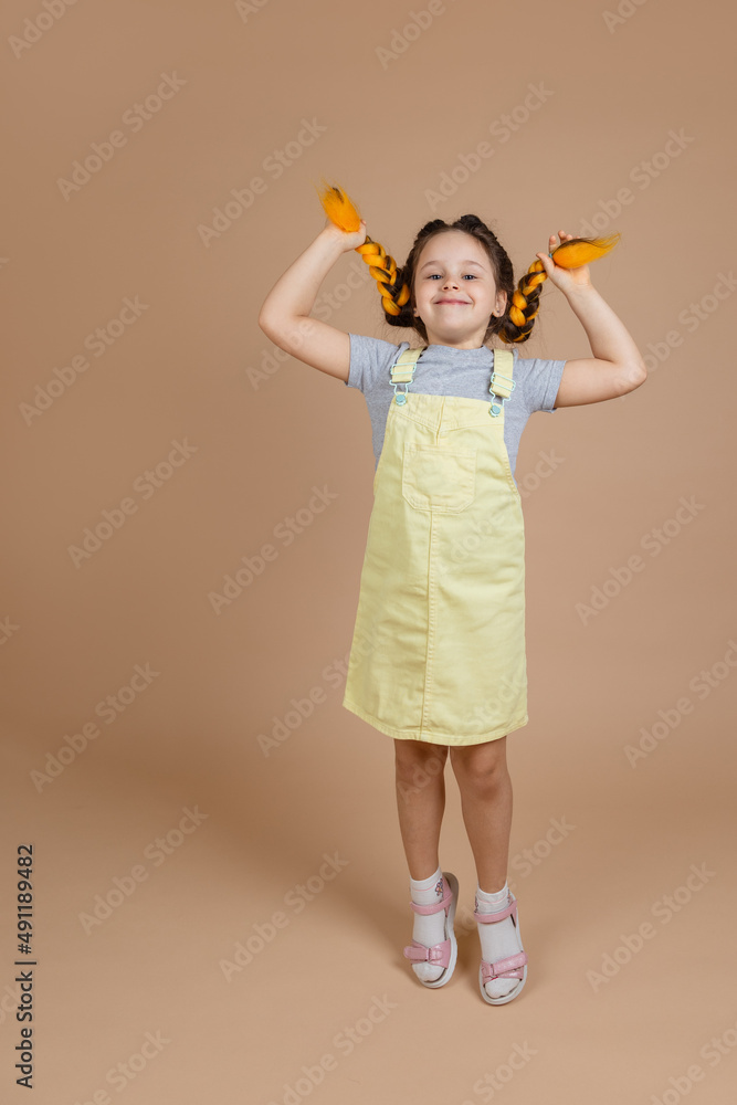 Cheerful small girl touching yellow kanekalon braids with hands, jumping and playing looking at camera with smile on face wearing yellow jumpsuit and gray t-shirt on beige background. Cheerful small girl touching yellow kanekalon braids with hands, jumping and playing looking at camera with smile on face wearing yellow jumpsuit and gray t-shirt on beige background.