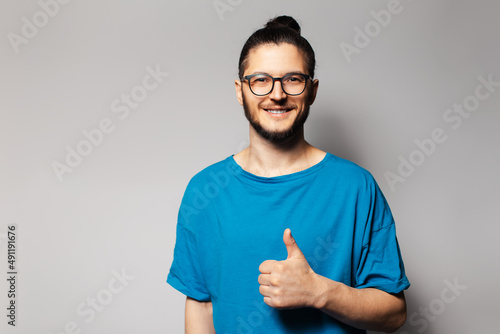 Studio portrait of young smiling man in blue shirt showing thumb up, on grey.