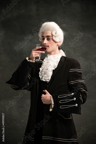 Handsome man in image of medieval person, aristocrat tasting wine isolated on dark green vintage background. Comparison of eras concept.