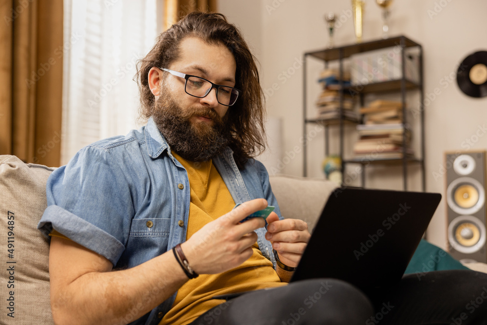 A bearded man is shopping at his favorite online store by paying with a credit card. He is sitting on a couch in the living room of a modern home. A bearded man is shopping at his favorite online store by paying with a credit card. He is sitting on a couch in the living room of a modern home.