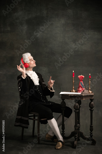 Fotografi Portrait of young man wearing wig and vintage medieval outfit like famous composer in modern headphones isolated on dark green vintage background