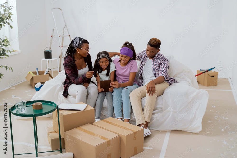 Joyful parents and their daughters are sitting on the couch in the apartment they just renovated, browsing room inspirations on the tablet while choosing paint colors for the walls Joyful parents and their daughters are sitting on the couch in the apartment they just renovated, browsing room inspirations on the tablet while choosing paint colors for the walls