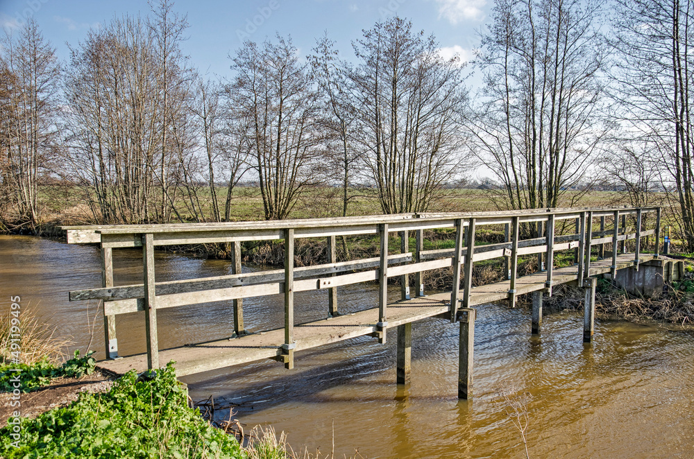 Narrow wooden pedestrian bridge across a wide ditch as part of a hiking trail on a sunny day in early spring in Krimpenerwaard region in the Netherlands Narrow wooden pedestrian bridge across a wide ditch as part of a hiking trail on a sunny day in early spring in Krimpenerwaard region in the Netherlands