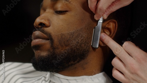 Man shaving beard of african american man with straight razor in barbershop