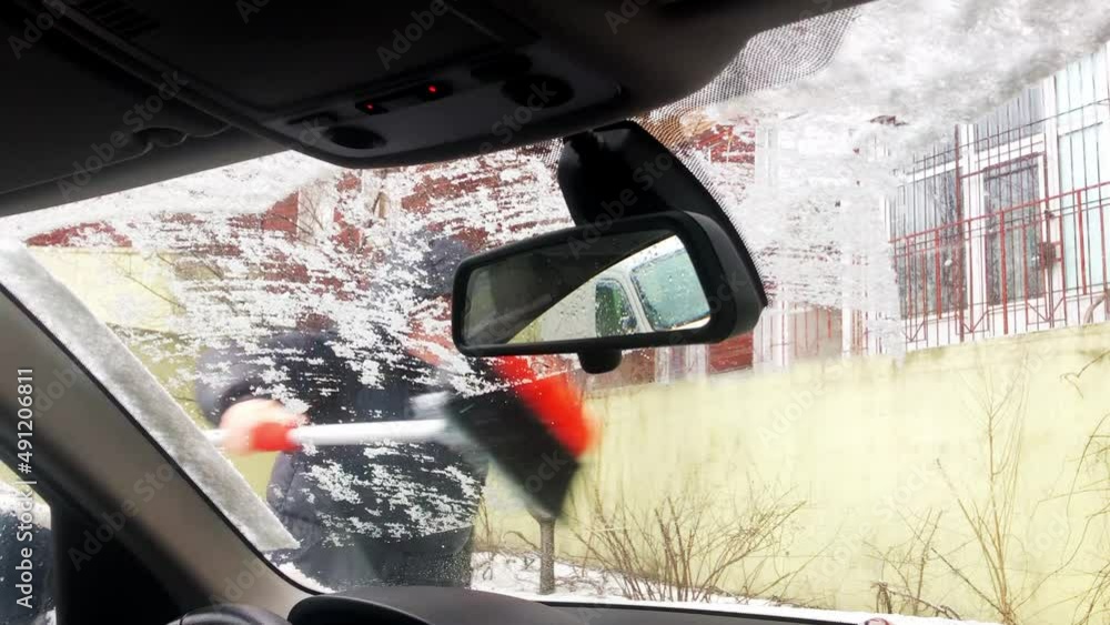 A woman cleaning car window with brush removing snow in frosty winter day. A driver preparing
