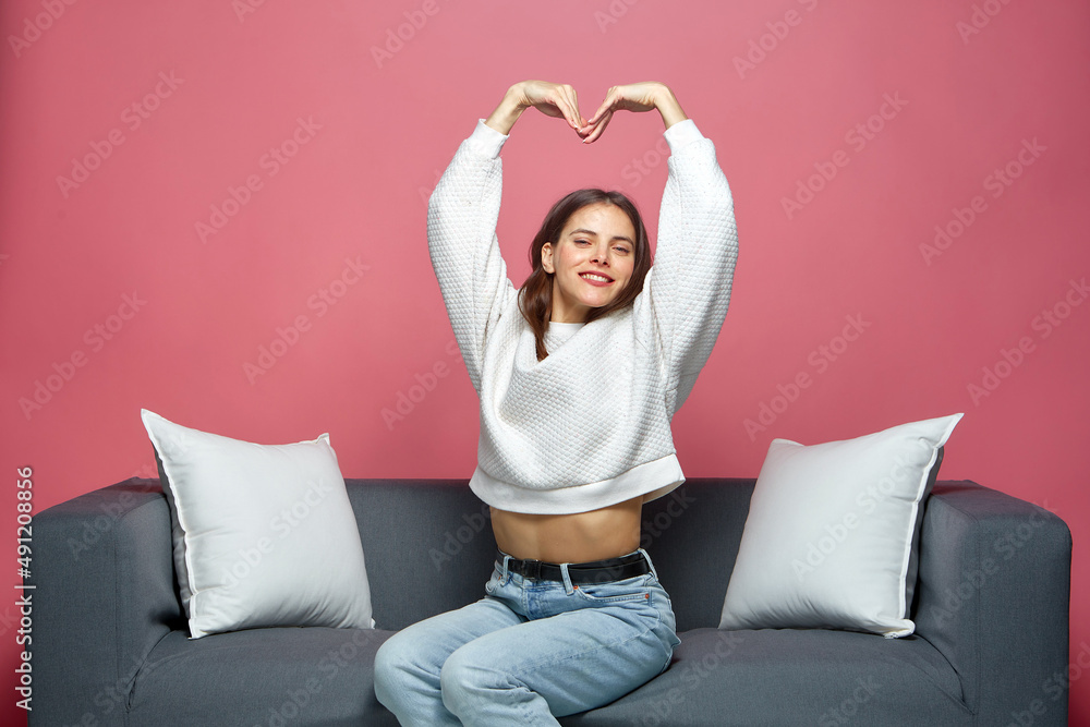 Smiling friendly young girl stretching hands up, showing heart, feeling love, sitting on sofa. Women wellness concept Smiling friendly young girl stretching hands up, showing heart, feeling love, sitting on sofa. Women wellness concept