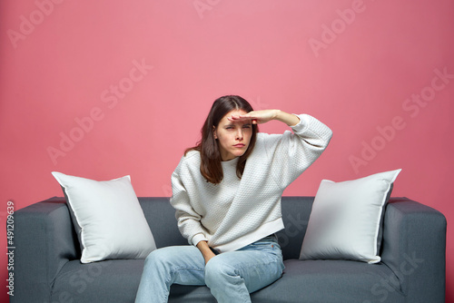 Attentive young girl looking forward with hand over forehead, sitting on sofa. Searching, future opportunities concept