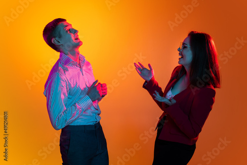 Portrait of two young people, man and woman, communicating in excitement isolated over orange studio background in neon