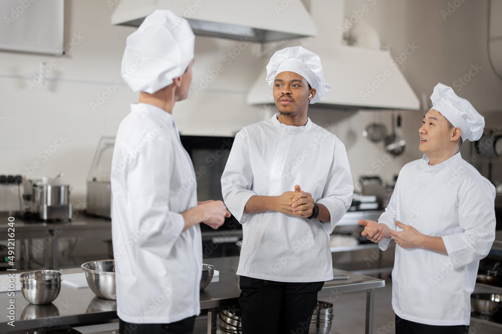 Three multiracial chef cooks talk while standing together during a break in the restaurant kitchen. Asian, Latin and European chefs wearing white uniform at work