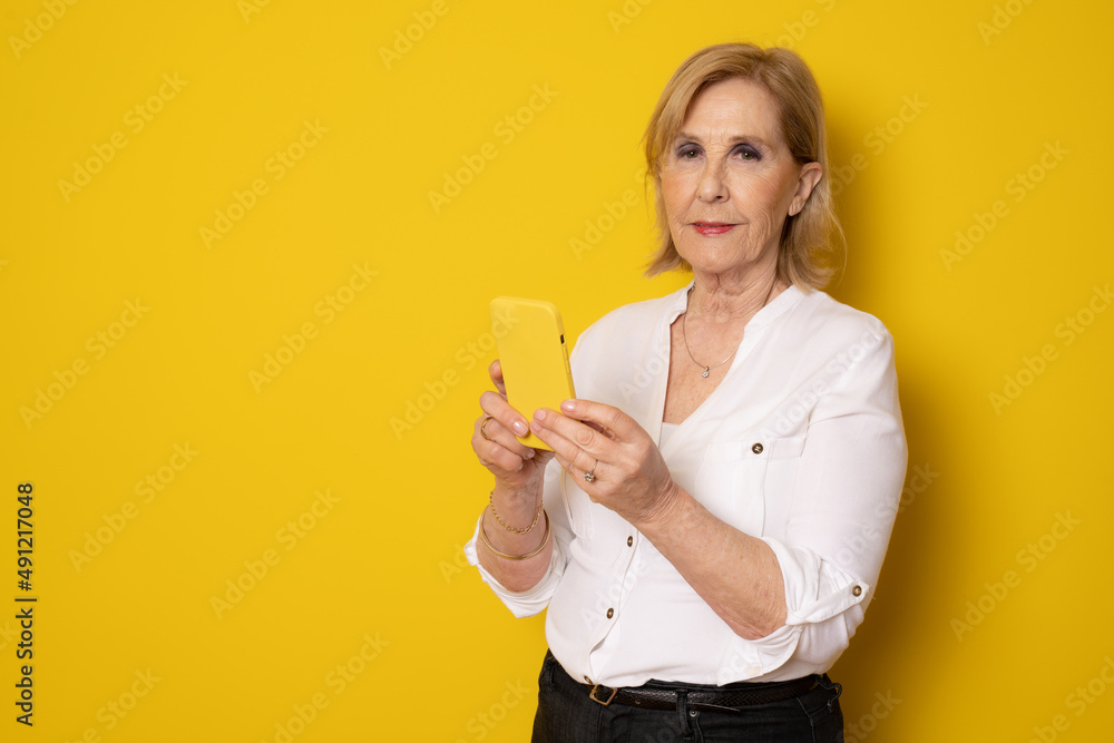 Happy mature senior woman using smartphone standing isolated over yellow background. Technology concept. Happy mature senior woman using smartphone standing isolated over yellow background. Technology concept.