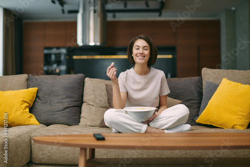 Smiling young girl with popcorn watching tv series or movie, sitting on sofa in living room at home, rest on weekend