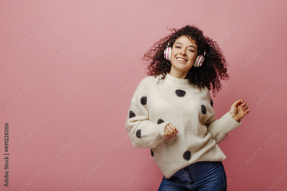 Smiling young caucasian lady is enjoying her favorite song and dancing on pink background. Portrait of curly brunette woman having fun in headphones. Audio technology, and music concept. Smiling young caucasian lady is enjoying her favorite song and dancing on pink background. Portrait of curly brunette woman having fun in headphones. Audio technology, and music concept.