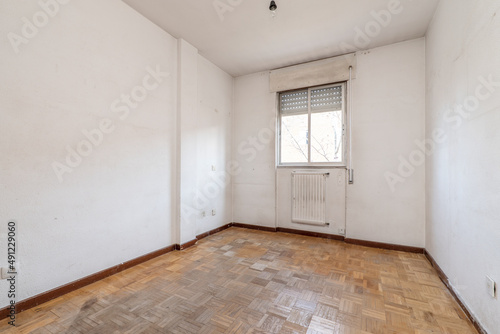 empty living room with white aluminum radiator under a window and dirty oak parquet flooring