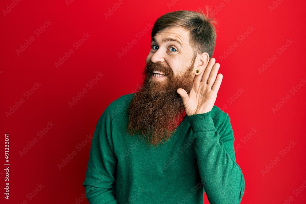 Redhead man with long beard wearing casual clothes smiling with hand over ear listening and hearing to rumor or gossip. deafness concept. Redhead man with long beard wearing casual clothes smiling with hand over ear listening and hearing to rumor or gossip. deafness concept.