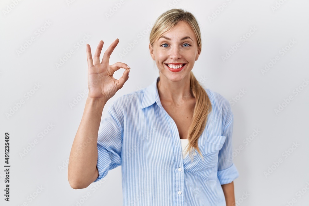 Fototapeta premium Beautiful blonde woman standing over white background smiling positive doing ok sign with hand and fingers. successful expression.