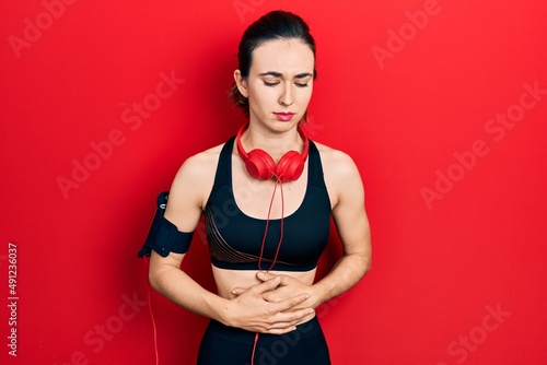 Young hispanic girl wearing gym clothes and using headphones with hand on stomach because indigestion, painful illness feeling unwell. ache concept.