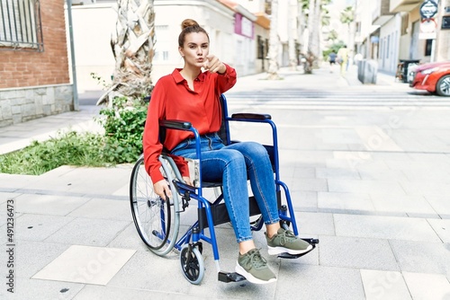 Young brunette woman sitting on wheelchair outdoors pointing with finger to the camera and to you, confident gesture looking serious
