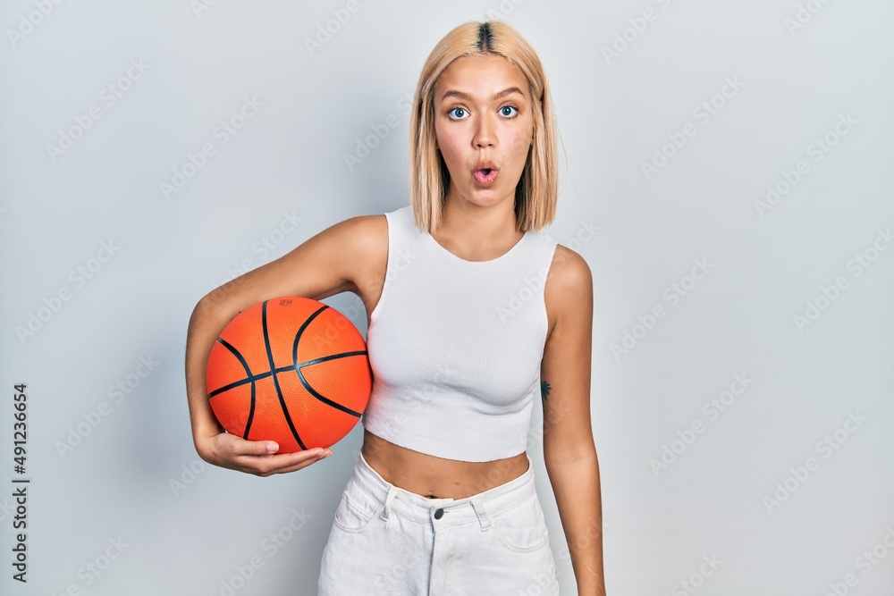 Beautiful blonde woman holding basketball ball scared and amazed with open mouth for surprise, disbelief face