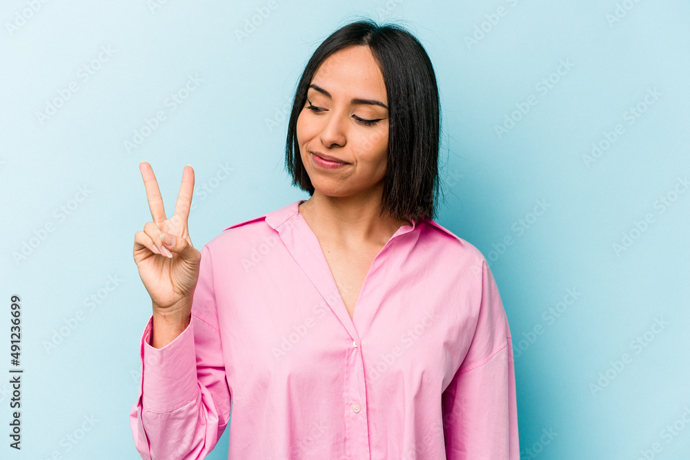 Young hispanic woman isolated on blue background joyful and carefree showing a peace symbol with fingers. Young hispanic woman isolated on blue background joyful and carefree showing a peace symbol with fingers.