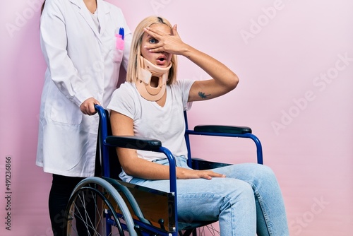 Beautiful blonde woman sitting on wheelchair with collar neck peeking in shock covering face and eyes with hand, looking through fingers with embarrassed expression.