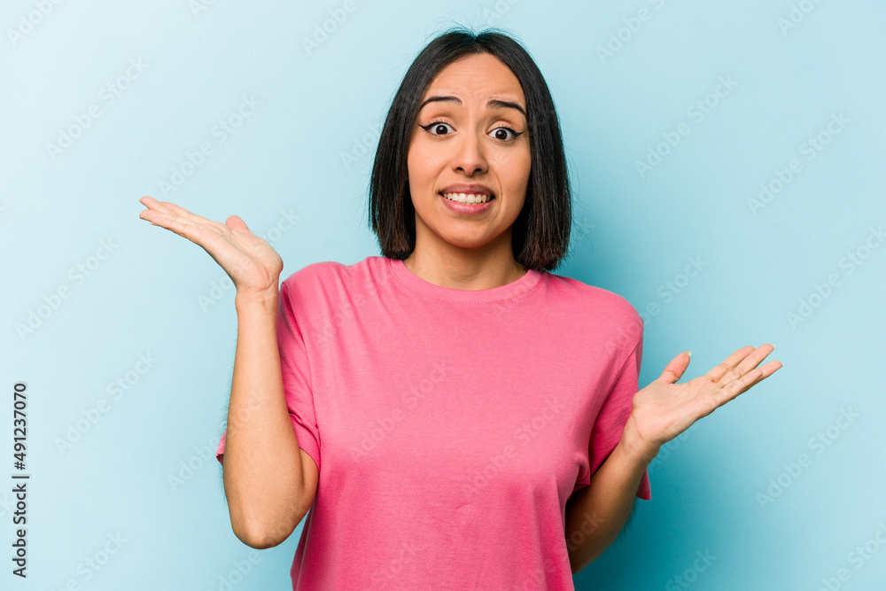 Young hispanic woman isolated on blue background confused and doubtful shrugging shoulders to hold a copy space. Young hispanic woman isolated on blue background confused and doubtful shrugging shoulders to hold a copy space.