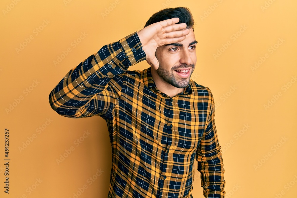 Young hispanic man wearing casual clothes very happy and smiling looking far away with hand over head. searching concept. Young hispanic man wearing casual clothes very happy and smiling looking far away with hand over head. searching concept.