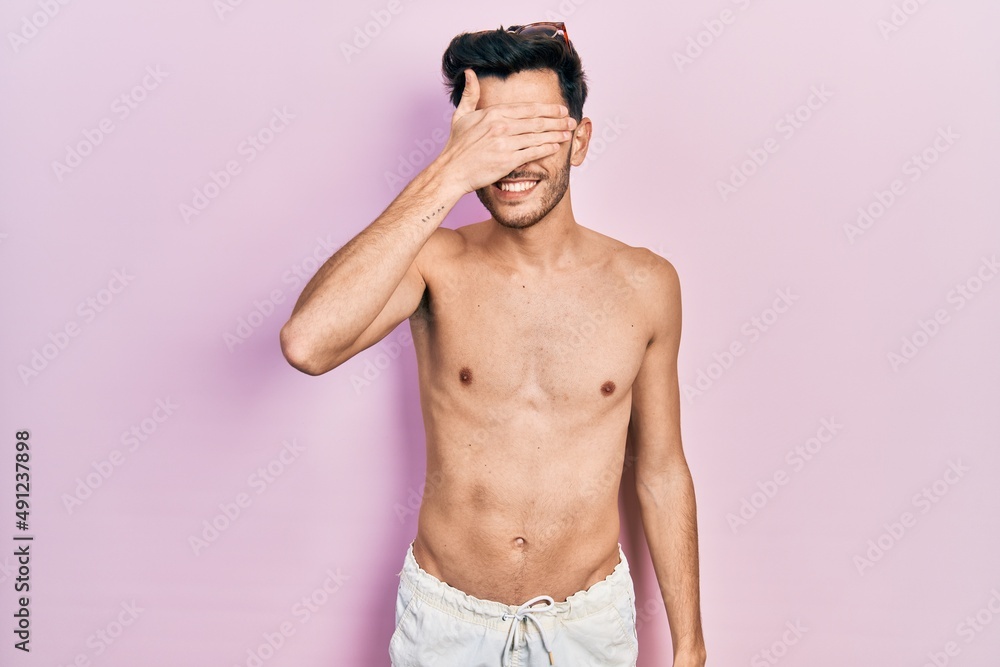 Fototapeta premium Young hispanic man wearing swimwear shirtless smiling and laughing with hand on face covering eyes for surprise. blind concept.